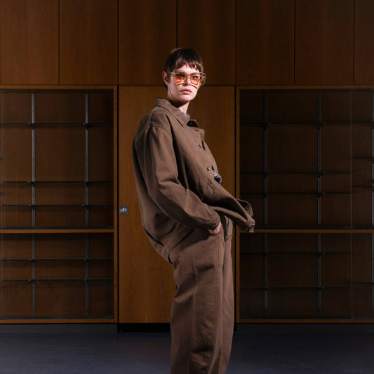 Person with short hair and tinted glasses wearing a loose brown outfit, standing in front of wooden cabinets with glass shelves.